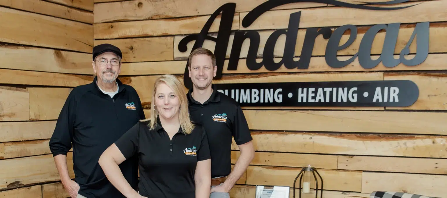 Three Andreas staff posing together in front of wood-paneled wall with Andreas logo in black.