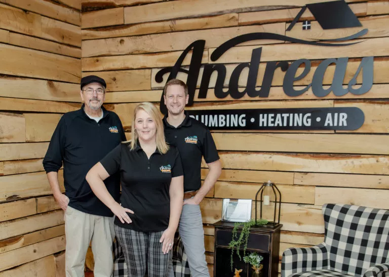 Three Andreas employees standing together next to large black Andreas logo on the wall behind them.
