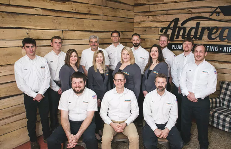 Team Andreas standing together in front of wood-paneled wall with black Andreas logo on it.