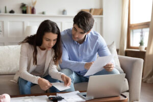 A couple sitting on a couch with a laptop and a calculator; they're looking through documents.