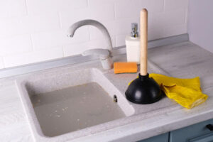 A clogged sink full of standing water with a plunger and gloves nearby on the countertop.