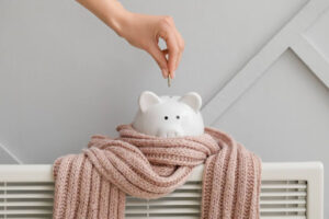 A person putting a coin into a piggy bank. The piggy bank is wrapped in a pink scarf and sitting on top of a radiator.